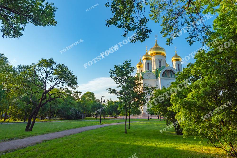 На фотографии изображен Екатерининский собор в Пушкине, окруженный зеленью парка. Величественное здание с золотыми куполами гармонирует с природным ландшафтом. Дорожки парка, ведущие к собору, создают атмосферу уюта и умиротворения. Летний солнечный день подчеркивает изящество архитектуры и спокойствие исторического места.